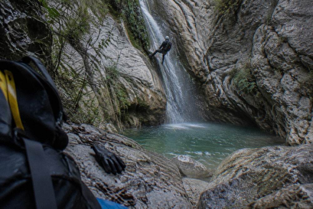 Canyoneer in a wetsuit rappelling beside a narrow waterfall into a green pool