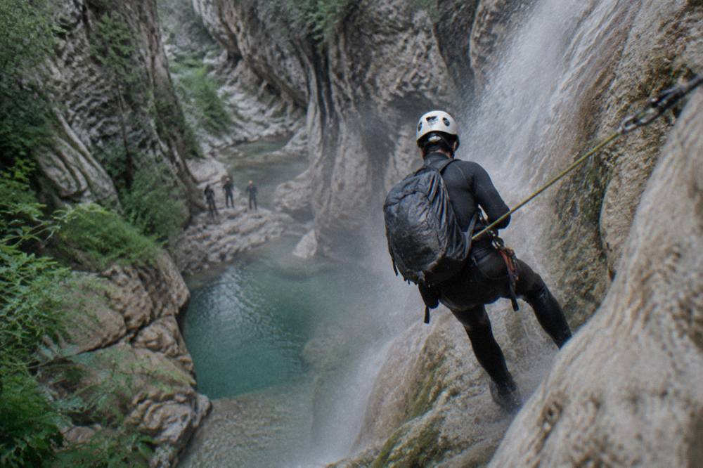 A canyoner in a wetsuit rappelling beside a waterfall into a pool below