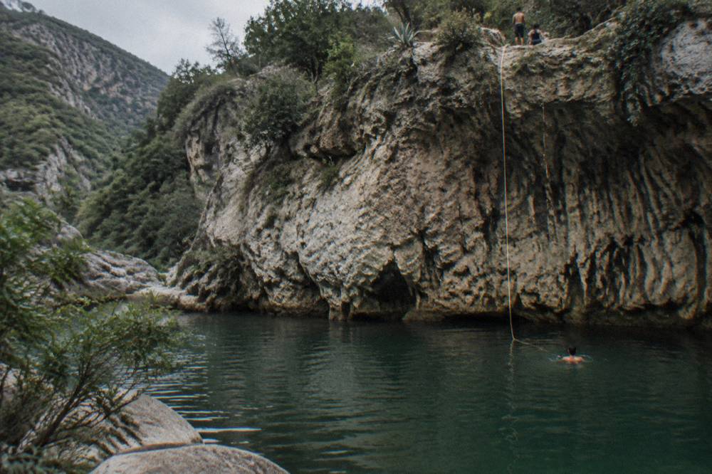 A deep green pool at the base of a canyon wall where a rope hangs from above