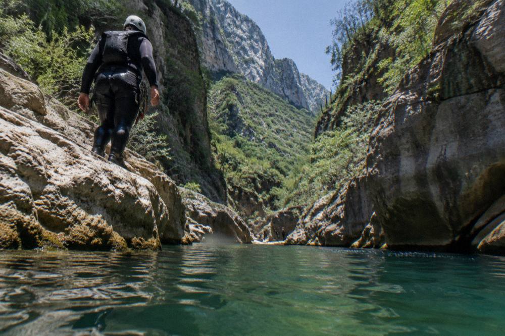 A canyoner in a wetsuit standing at the edge of a rock ledge above a deep green pool