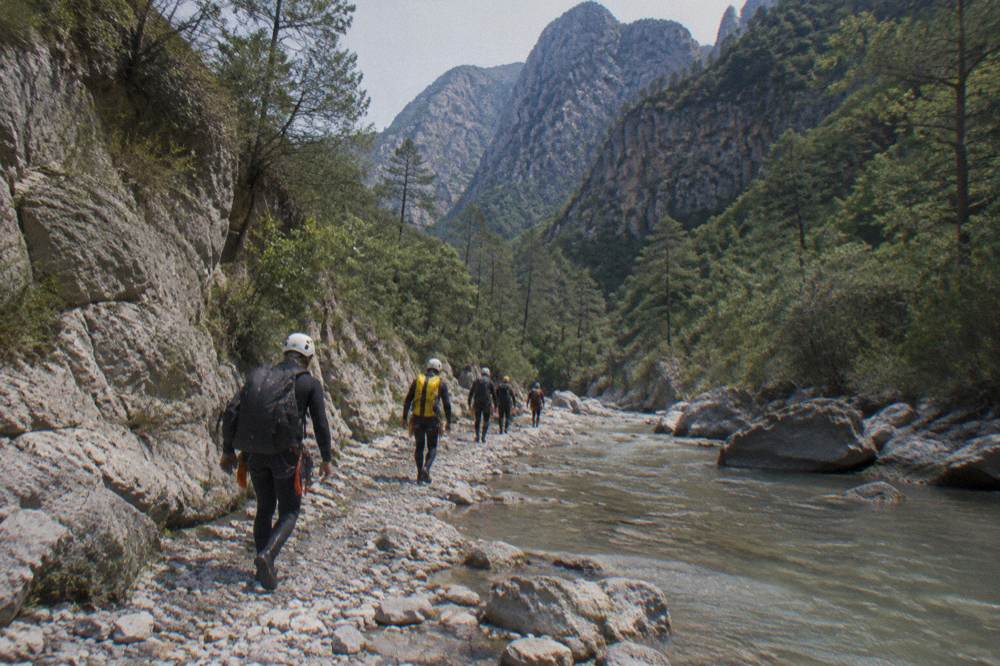 A line of canyoners in helmets and wetsuits walking along a rocky stream bed between canyon walls