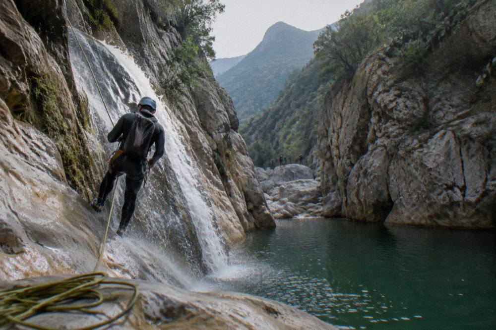 A canyoner in a wetsuit rappelling beside a narrow waterfall into a green pool
