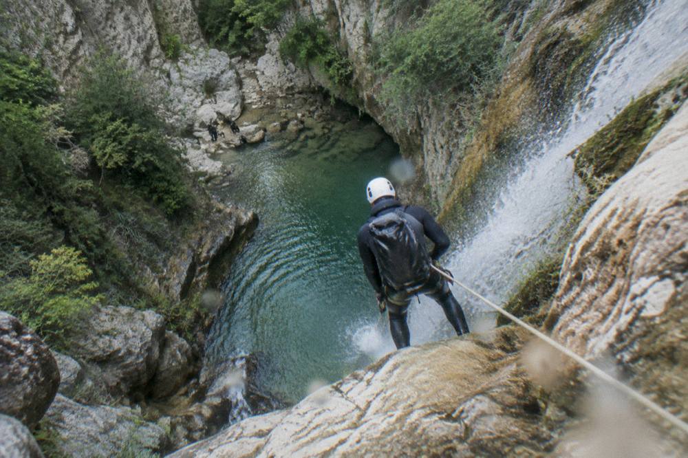 Canyoneer in a wetsuit rappelling beside a waterfall into a green pool inside a narrow canyon