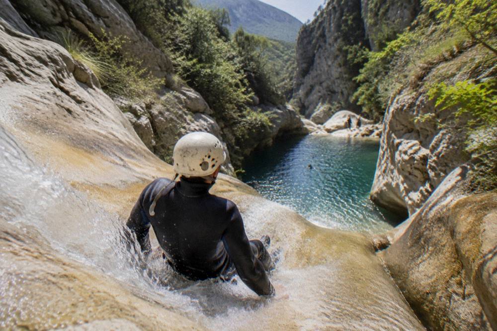Canyoneer sliding down a smooth rock chute into a deep blue pool framed by tall canyon walls