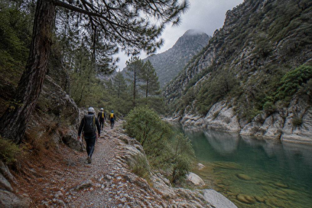 Group of canyoneers in helmets and wetsuits walking a rocky trail beside a calm green river in a pine forest