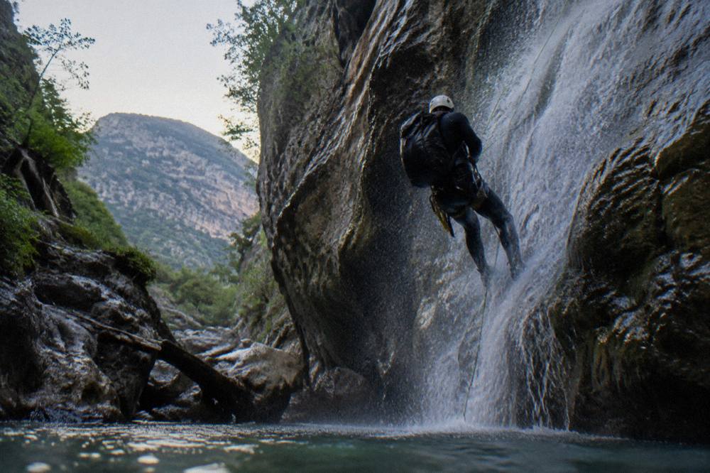 Canyoneer in a wetsuit rappelling beside a narrow waterfall inside a shaded slot canyon