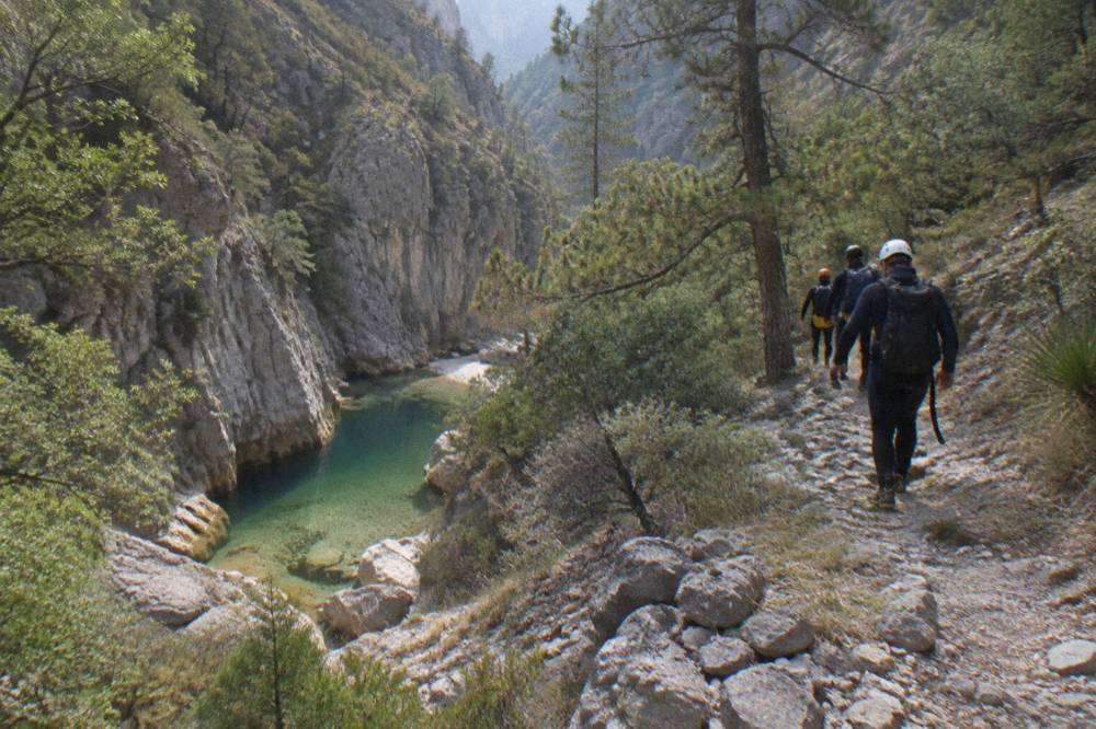 Group of canyoners in helmets and wetsuits walking a rocky trail above a green canyon pool