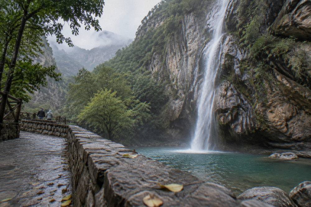 A narrow waterfall falling down a tall rock face into a pool surrounded by trees