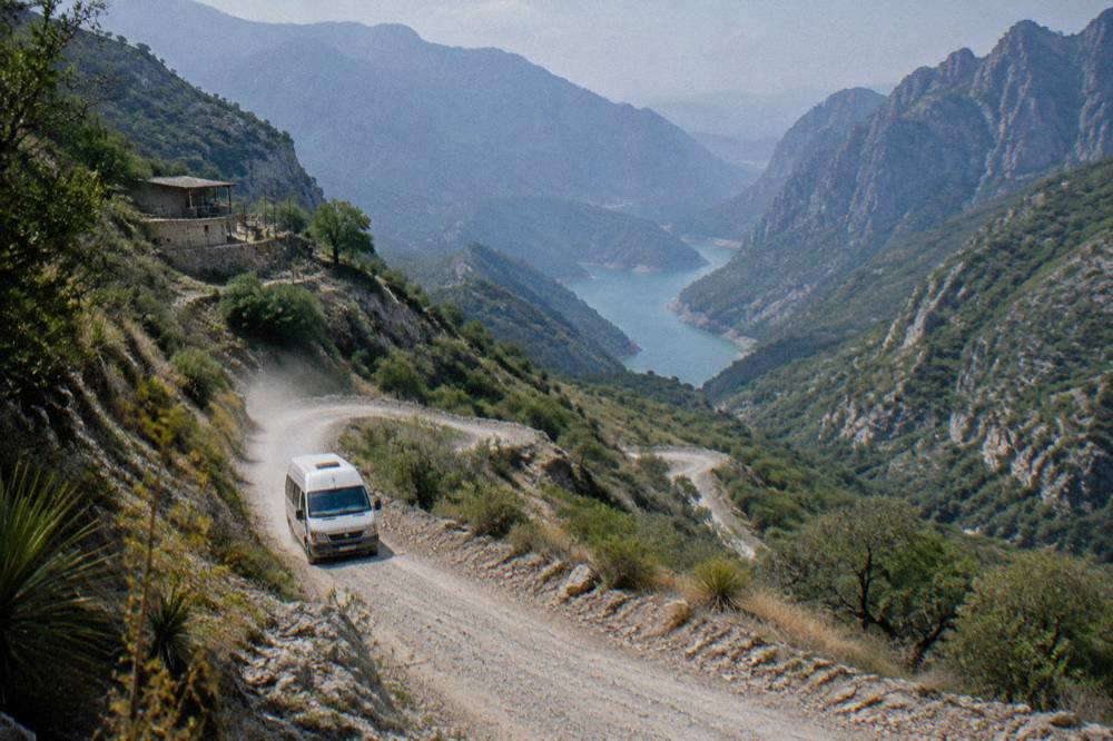 A winding mountain road through green hills with a reservoir in the distance