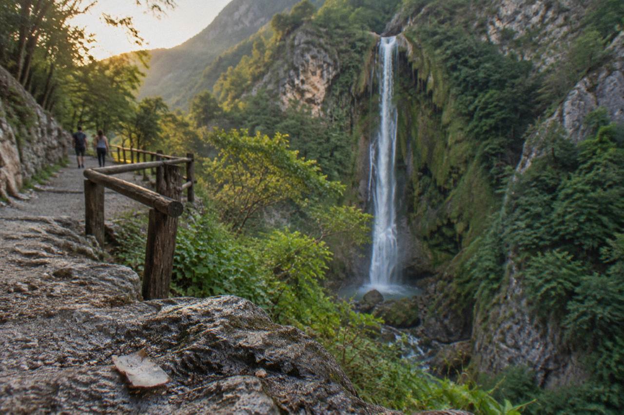 A tall thin waterfall dropping over a green cliff into a forested canyon