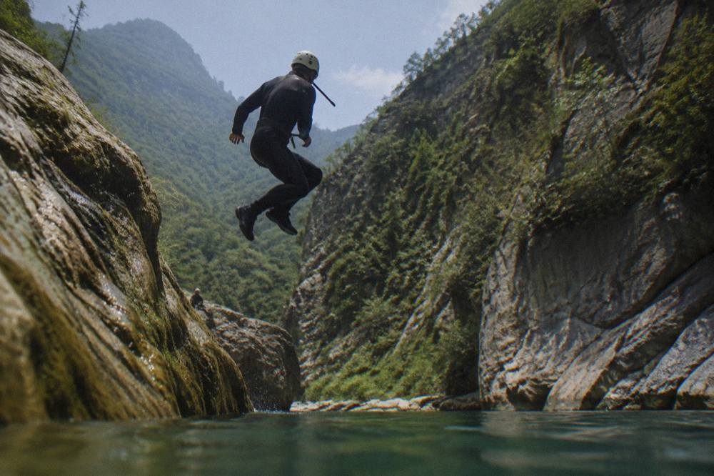 Canyoneer in a wetsuit mid-jump from a rock ledge into a deep green pool