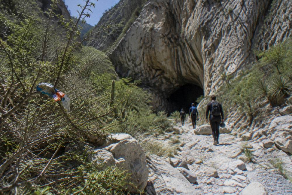 Group of hikers in wetsuits walking a rocky canyon trail toward a dark cave entrance