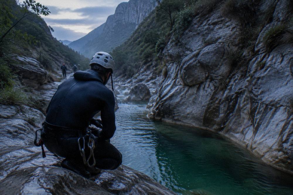 Canyoneer in a wetsuit and helmet preparing to jump from a rock ledge into a green pool below
