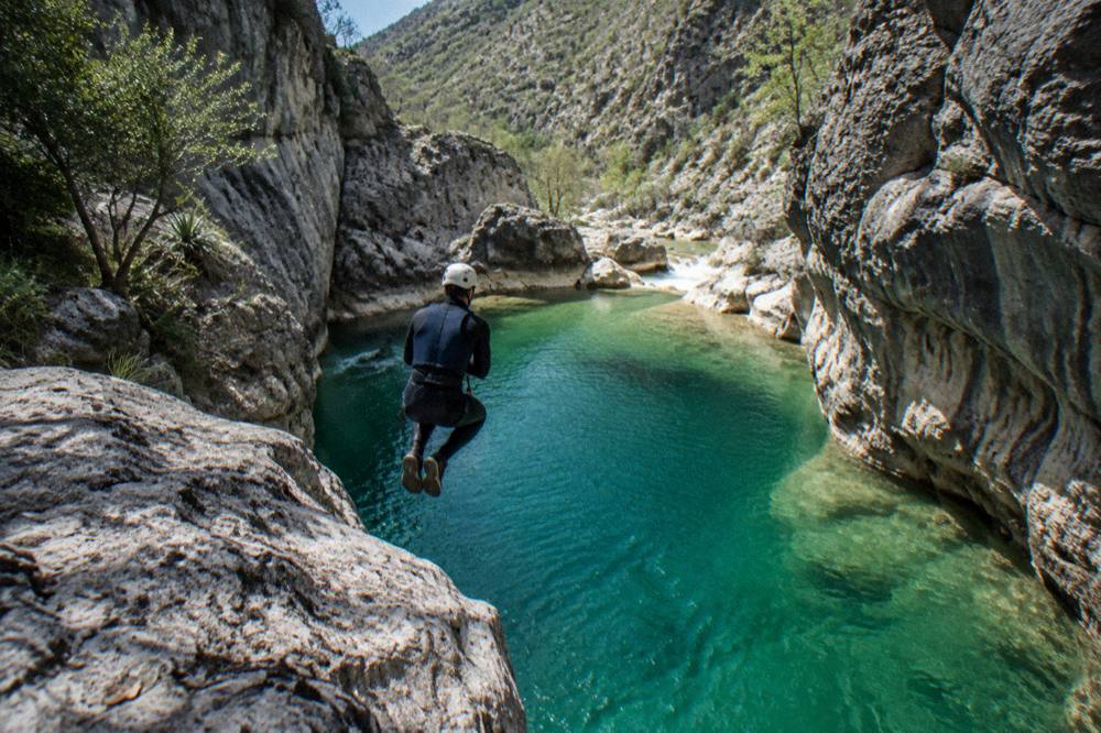Canyoneer mid-jump from a rocky ledge into a deep turquoise pool