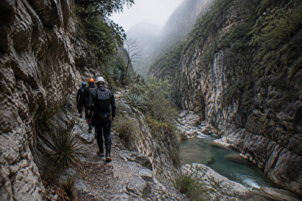 Group of hikers in wetsuits and helmets walking a canyon trail between rock walls