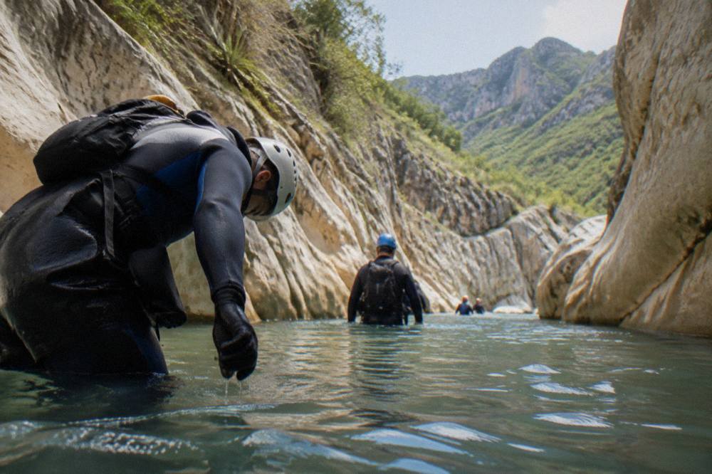 Canyoneers in wetsuits and helmets wading through a waist-deep pool between rock walls