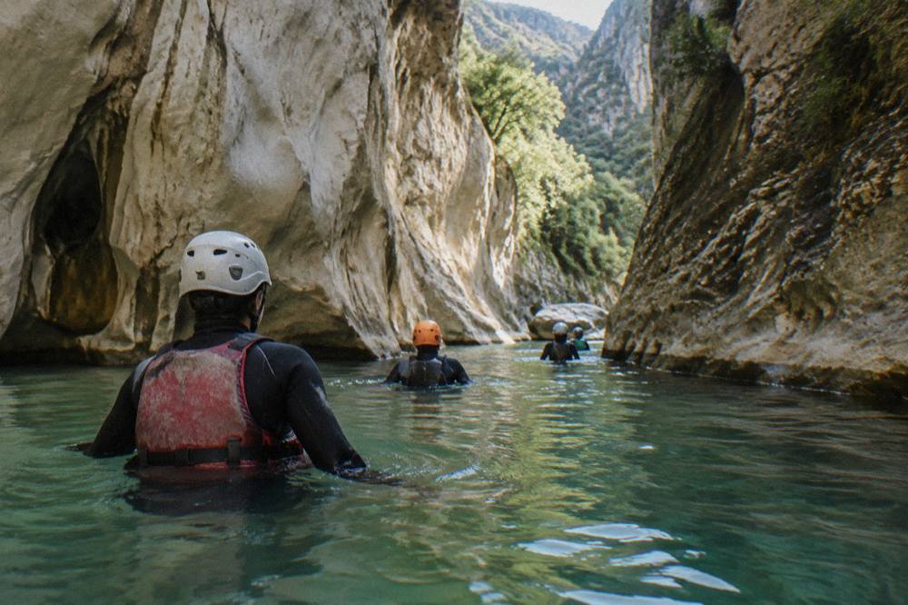 Canyoneers in wetsuits and helmets wading through a green pool between canyon walls