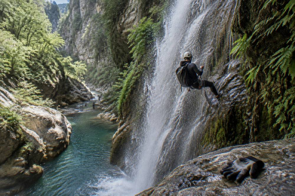 A canyoner mid-rappel beside a tall waterfall in a green gorge