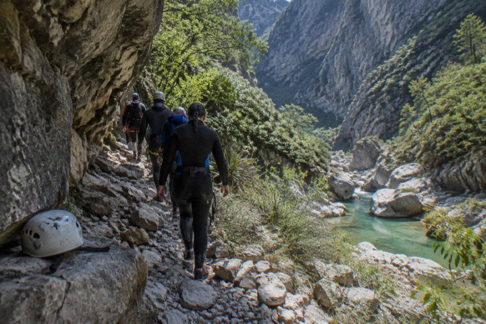 Canyoneers in wetsuits and helmets walking a rocky trail beside a canyon wall