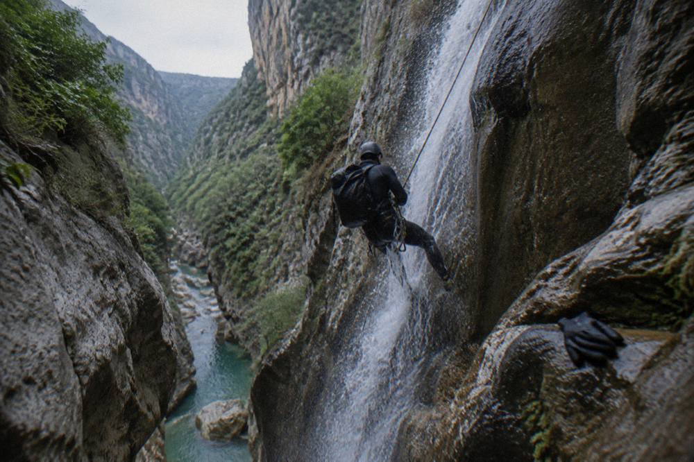 A canyoner in a wetsuit rappelling beside a narrow waterfall between dark rock walls