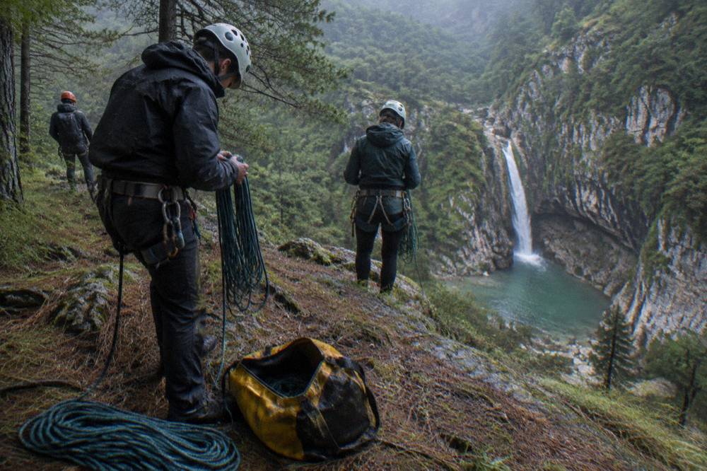 A guided group in helmets and harnesses preparing ropes at the lip of a forested canyon rappel